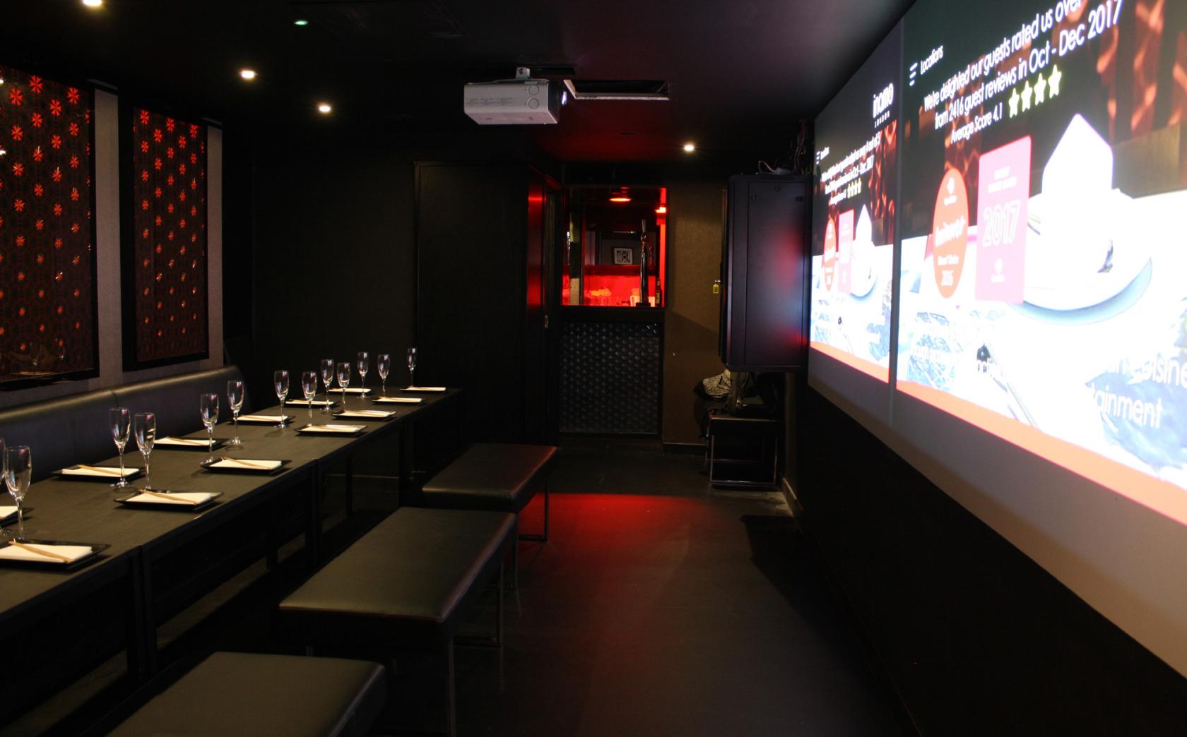 A restaurant with long black benches, set tables with glassware, and a wall projector screen at Games Room.