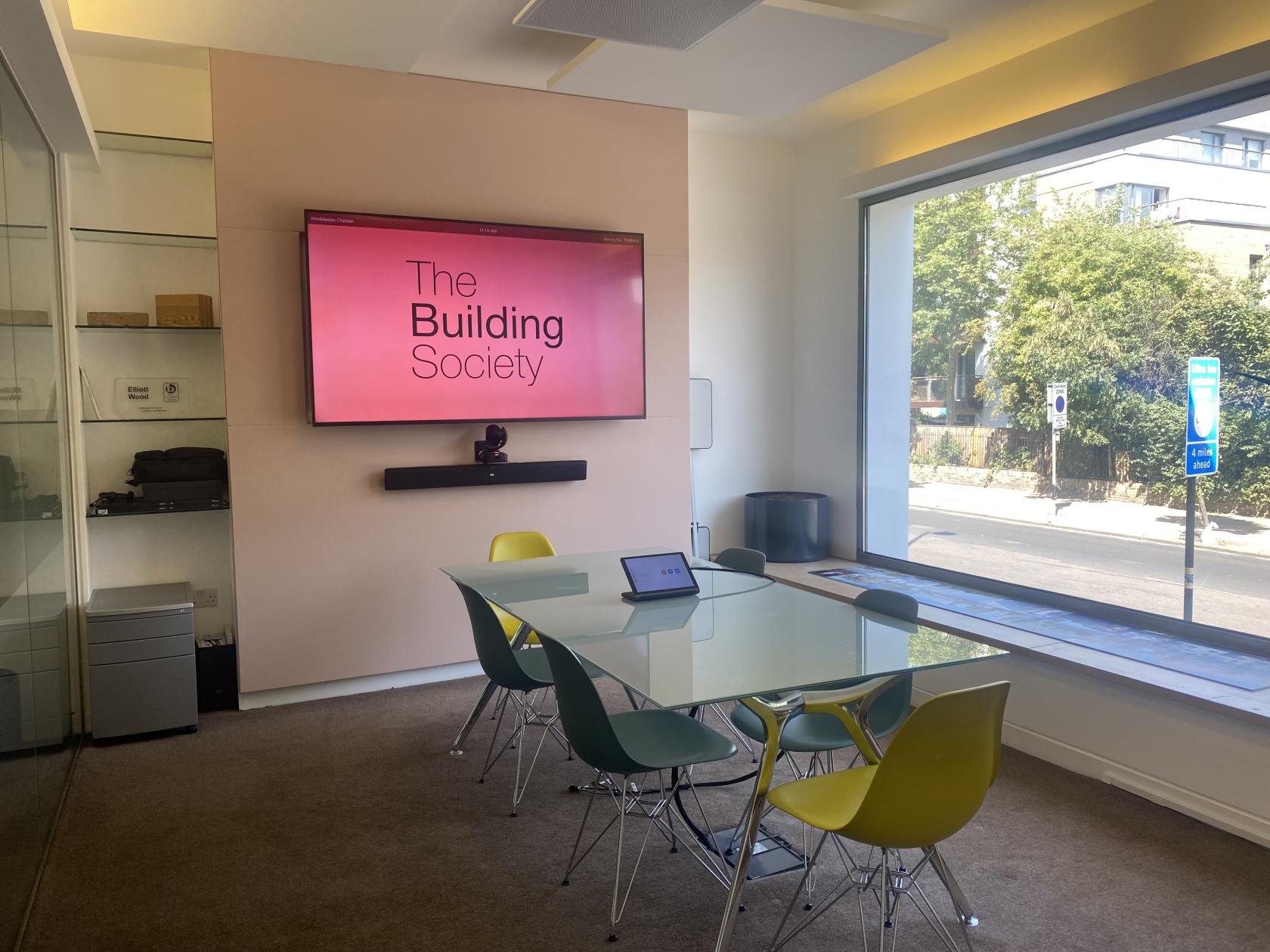 A meeting room with a glass table, colorful chairs, large window, and wall-mounted screen at The Society Building - Wimbledon.