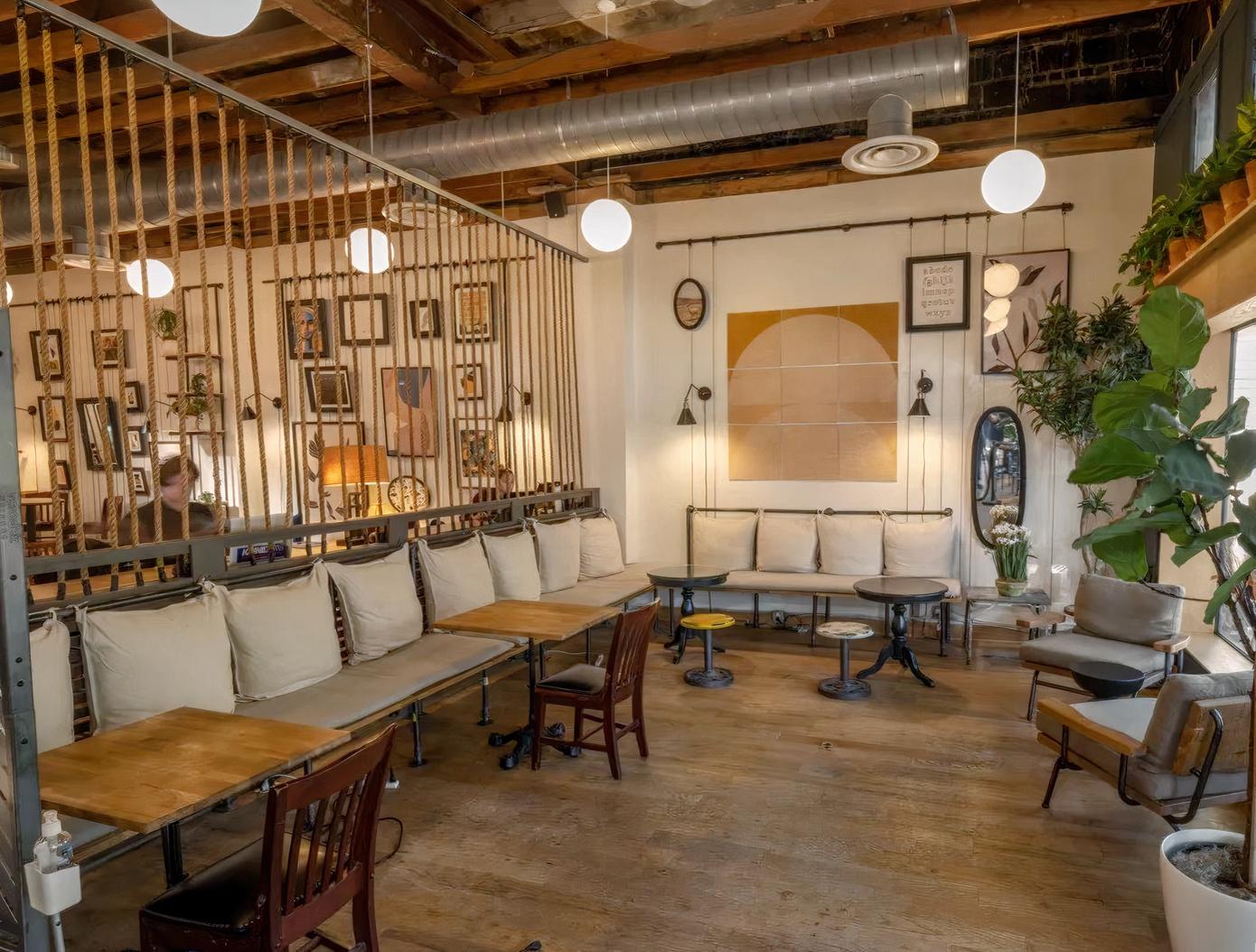 A cafe with wooden floors, exposed ceiling pipes, cushioned bench seating, and framed art at Three Whistles - Eat Drink Work.