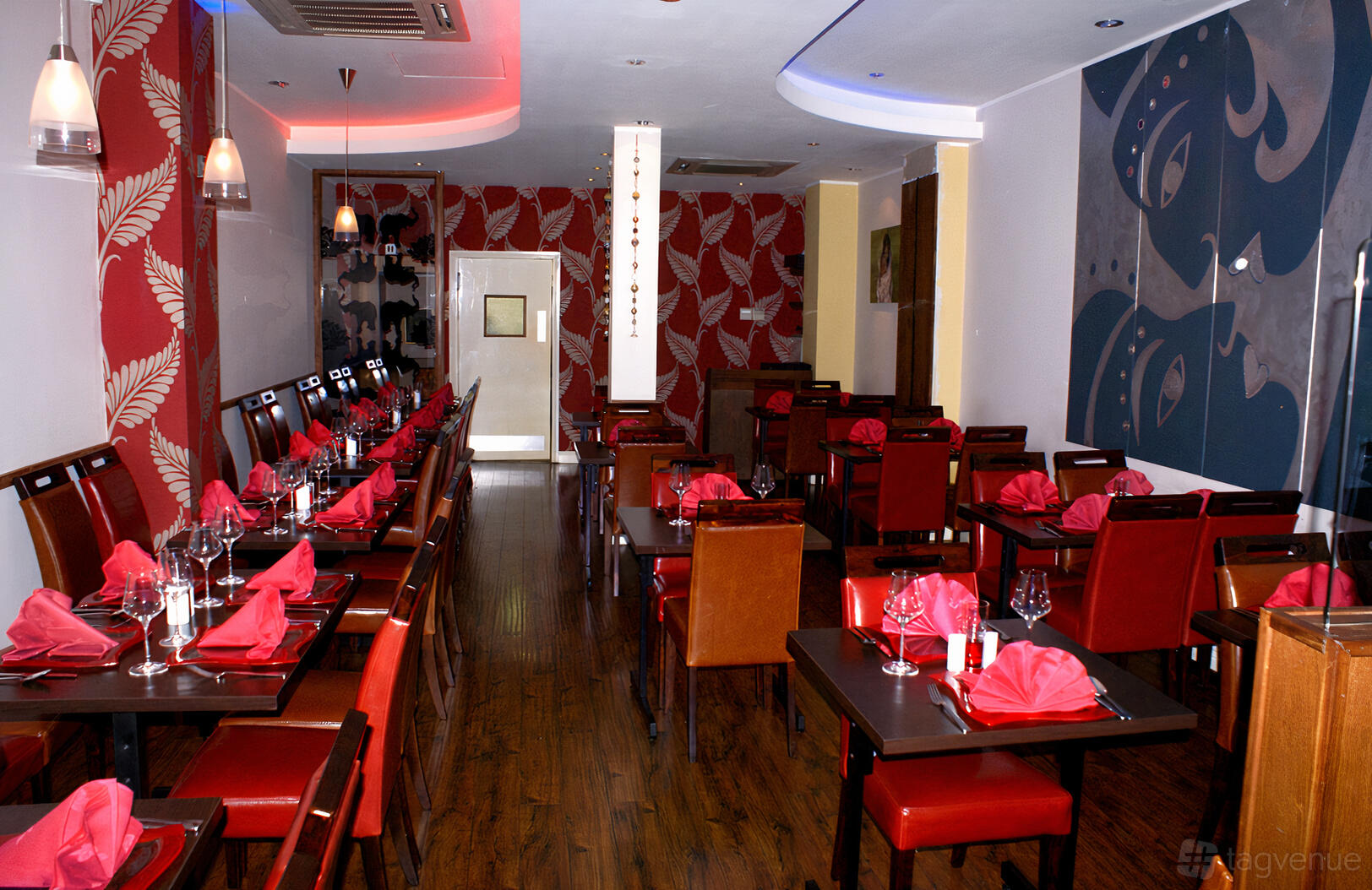 A restaurant dining area with red napkins on dark tables, wooden floors, and patterned accent walls at Shad Indian Restaurant.