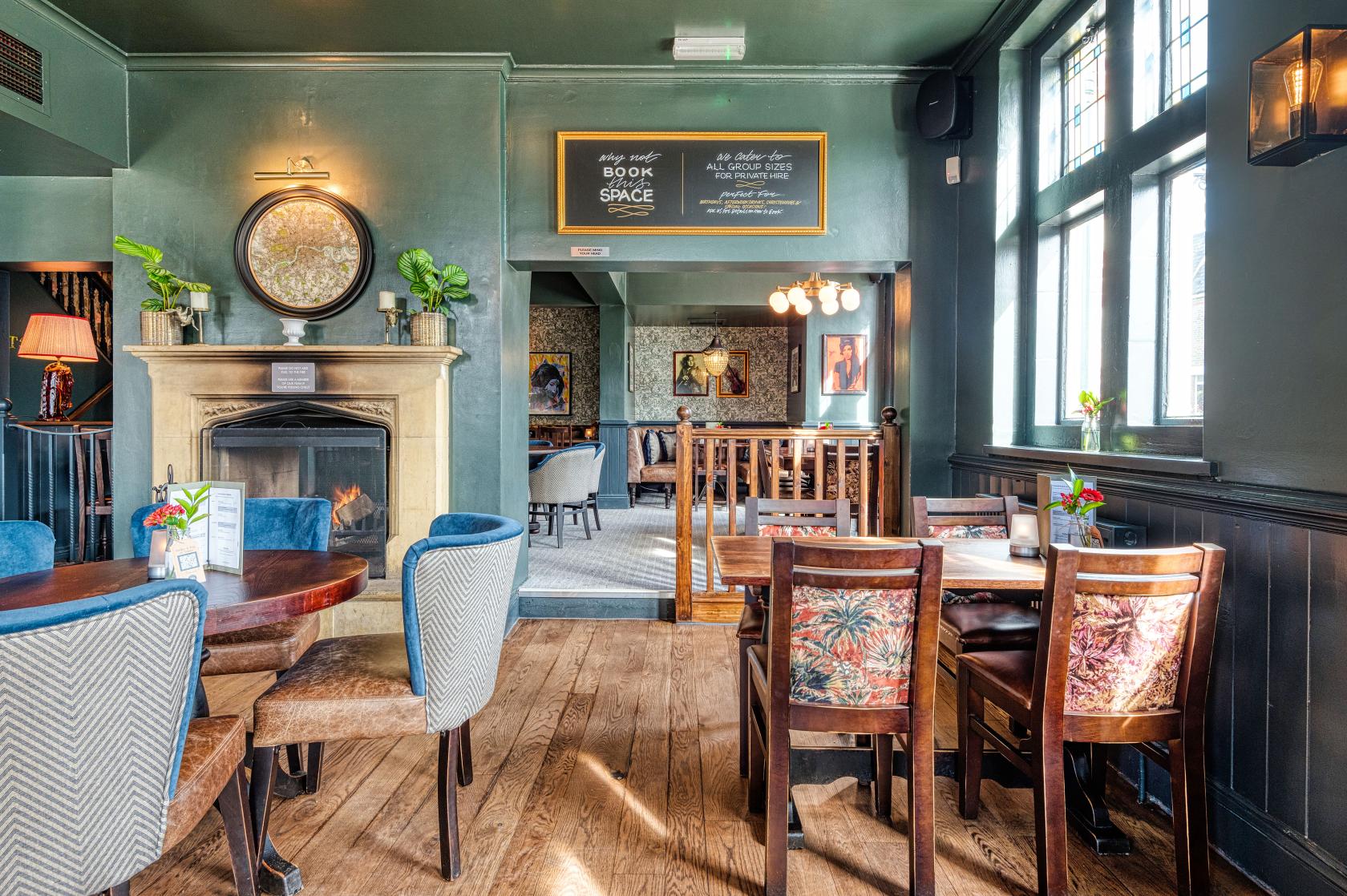 A pub function room with wooden floors, a fireplace, mix of armchairs and dining chairs, and large windows at The Salisbury Arms Winchmore Hill.