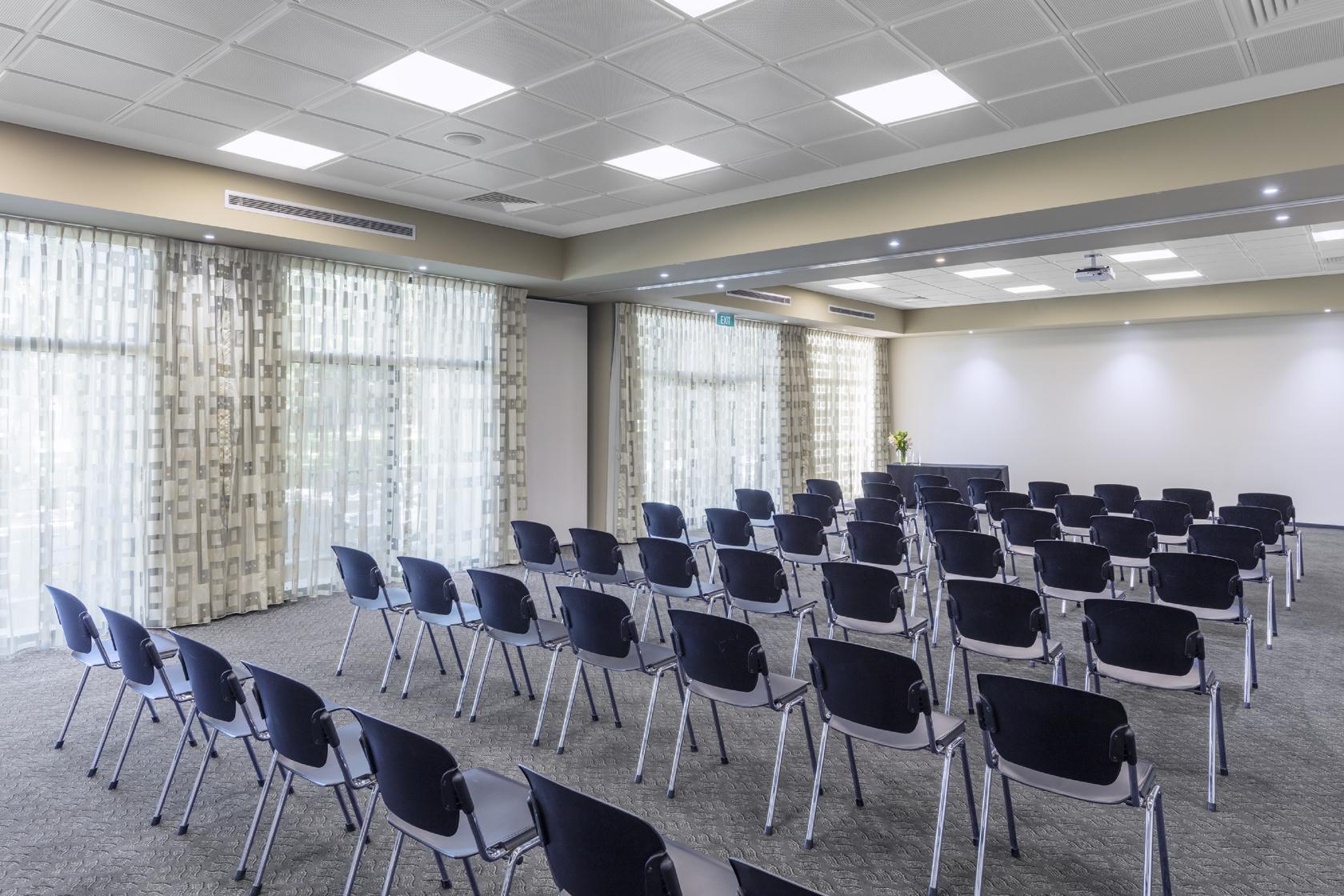 An apartment event space with rows of black chairs, carpeted floors, and large windows with sheer curtains at Nesuto Mounts Bay Perth Apartment Hotel.