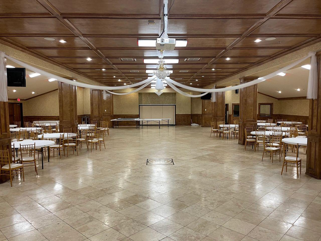 A spacious function room with tile flooring, round tables with chairs, draped ceiling fabric, and chandeliers at Victoria Folake Event Center.