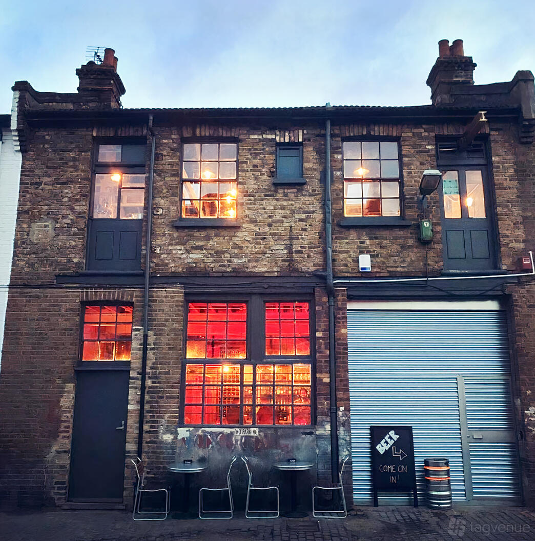 A pub with an exposed brick facade, large industrial windows, and warm glowing interior lights at Wolfpack Queens Park.