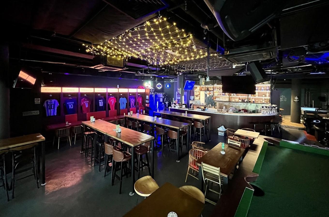 A sports bar with jerseys on the wall, string lights overhead, and high tables with stools at Bar Soccer.