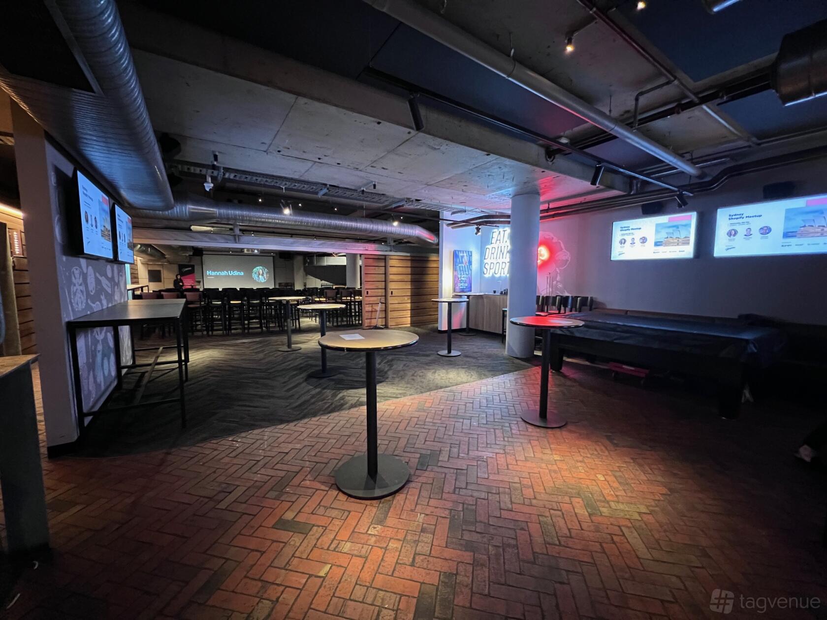 An event space in a pub with high tables, exposed ceilings, digital screens, and brick flooring at The Sporting Globe King Street Wharf.