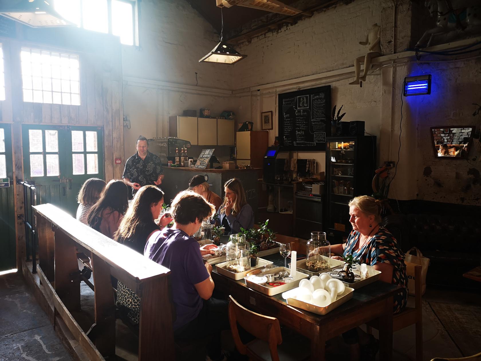 A cafe with communal wooden tables, exposed brick walls, large windows, and plants on tables at Artefact.