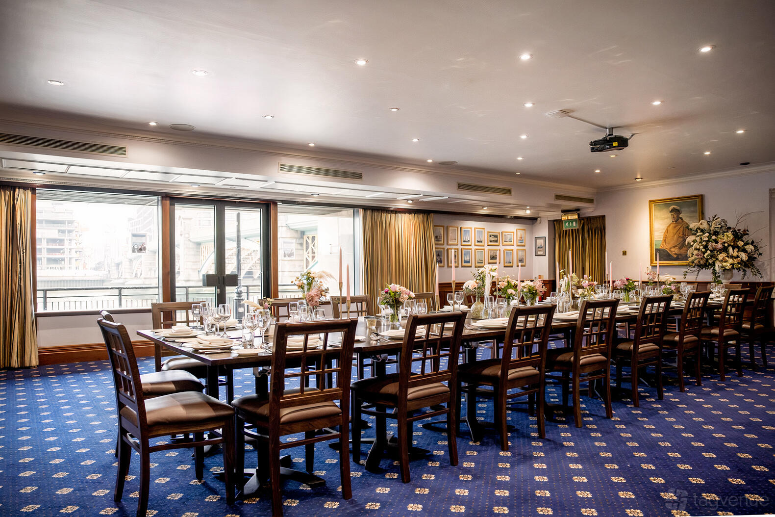 A dining room with long banquet tables, blue patterned carpet, wall portraits, and large windows at Little Ship Club.