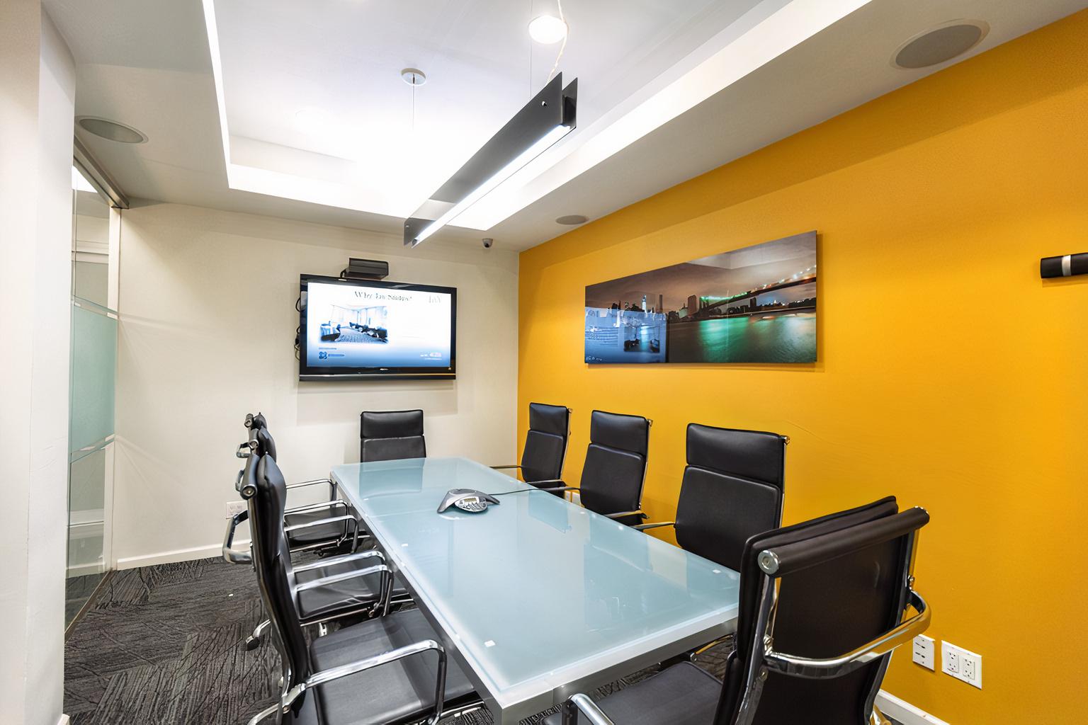 A meeting room with a glass conference table, black leather chairs, and a wall-mounted screen at Grand Central Jay Suites.