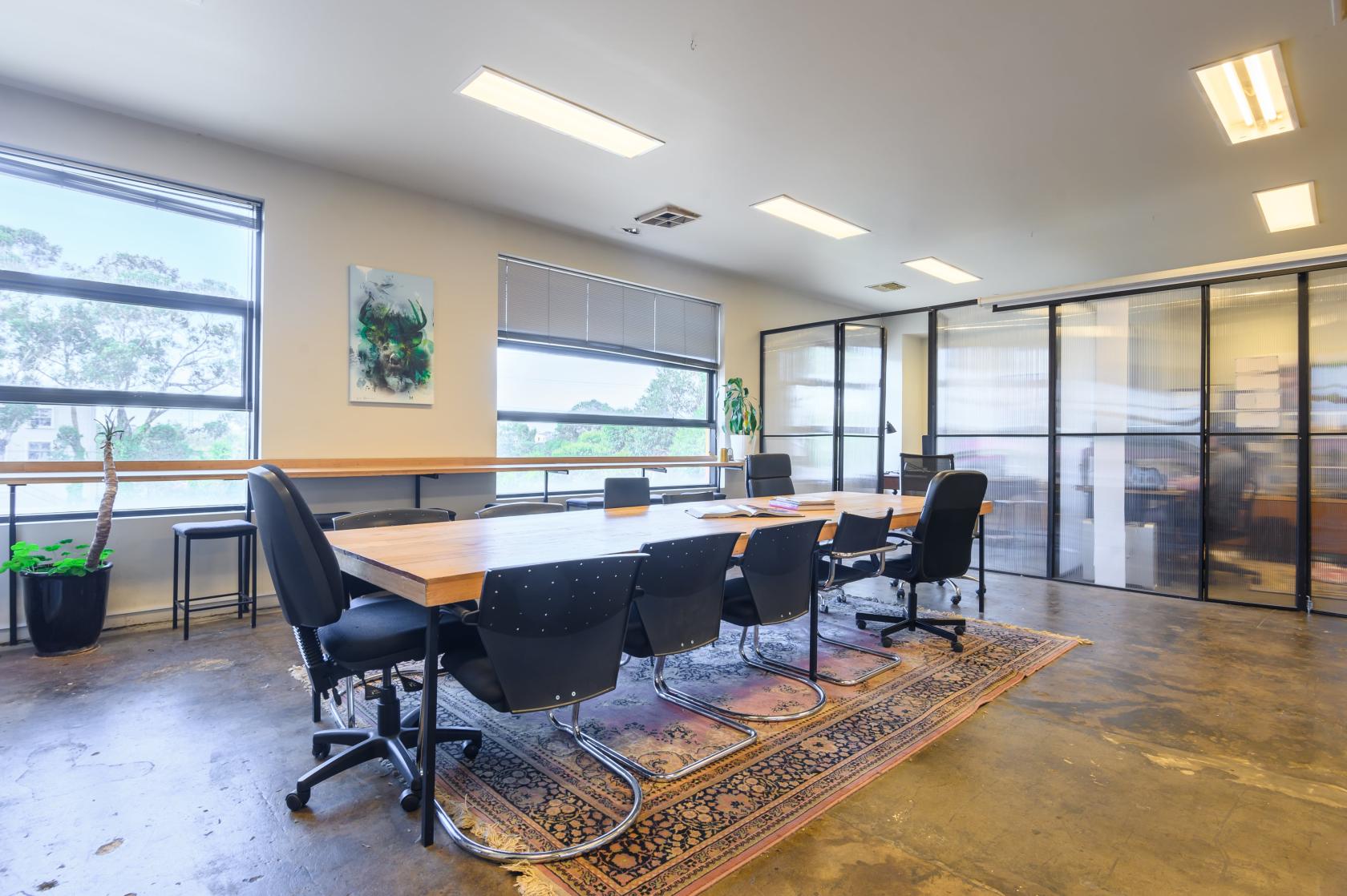 A coworking space with a wooden table, black chairs, floor-to-ceiling windows, and potted plants at Mycelium Studios.