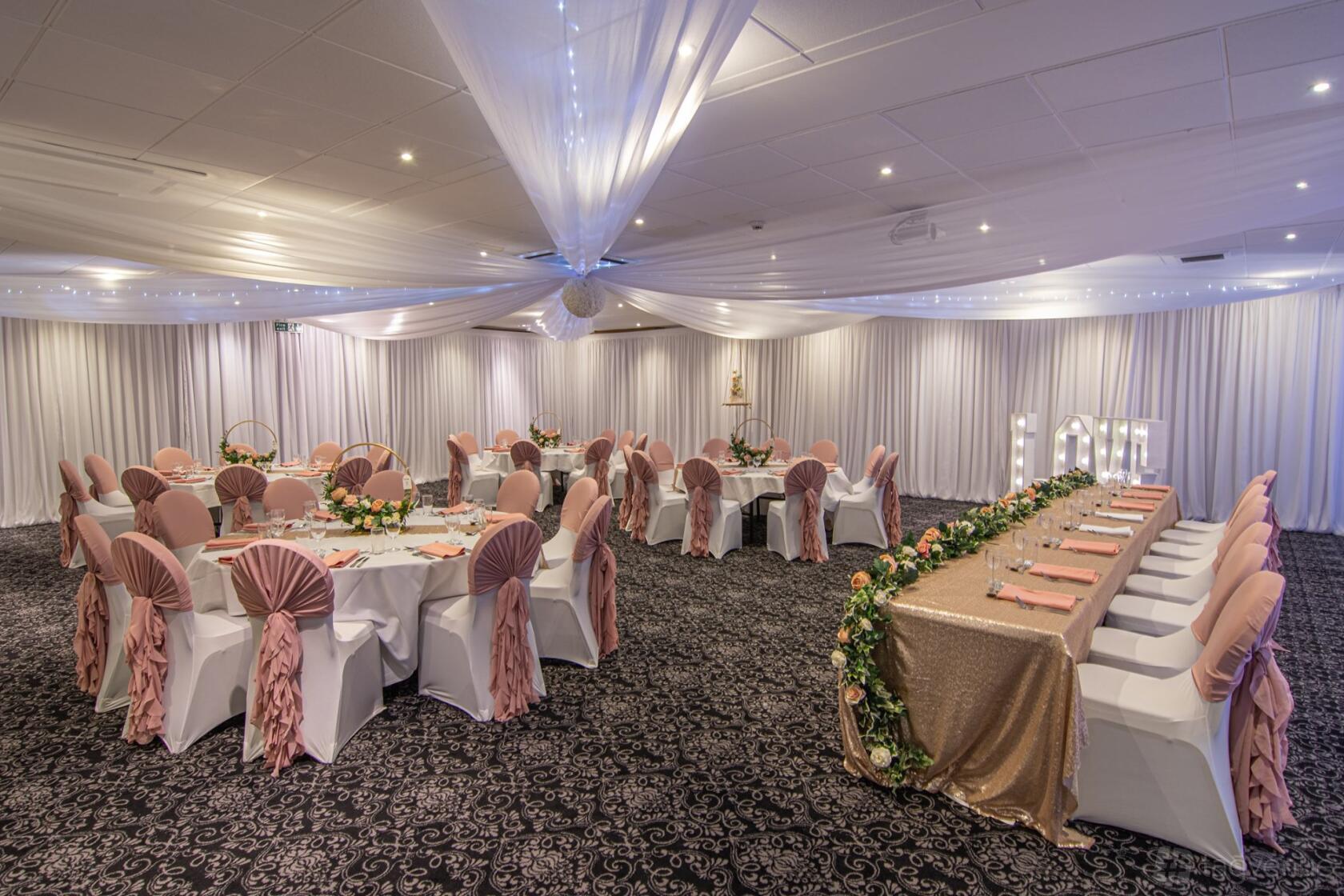 An event space with draped white ceilings, round tables and chairs with blush covers at Holiday Inn Corby Heart Suite.