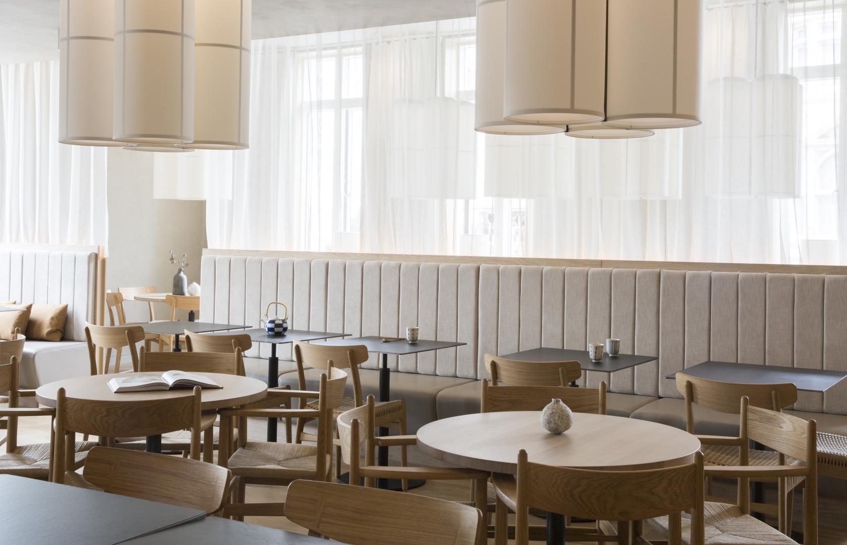 A restaurant with light wood tables, upholstered banquette seating, and tall drum pendant lights at Sticks'n'Sushi Chelsea.
