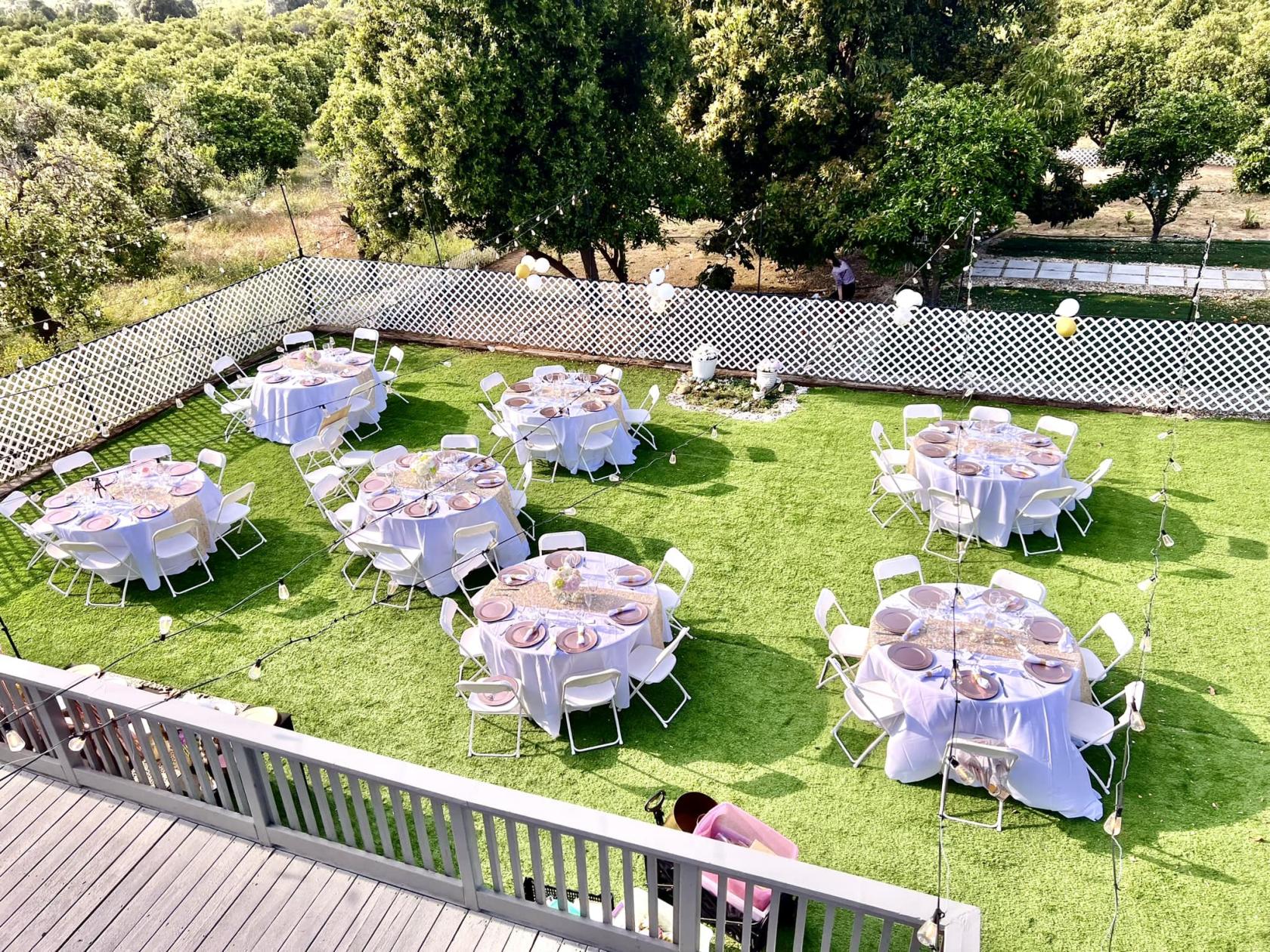 An outdoor event venue with round tables draped in white linens on a lawn, bordered by white lattice fencing at King Street Orchard.