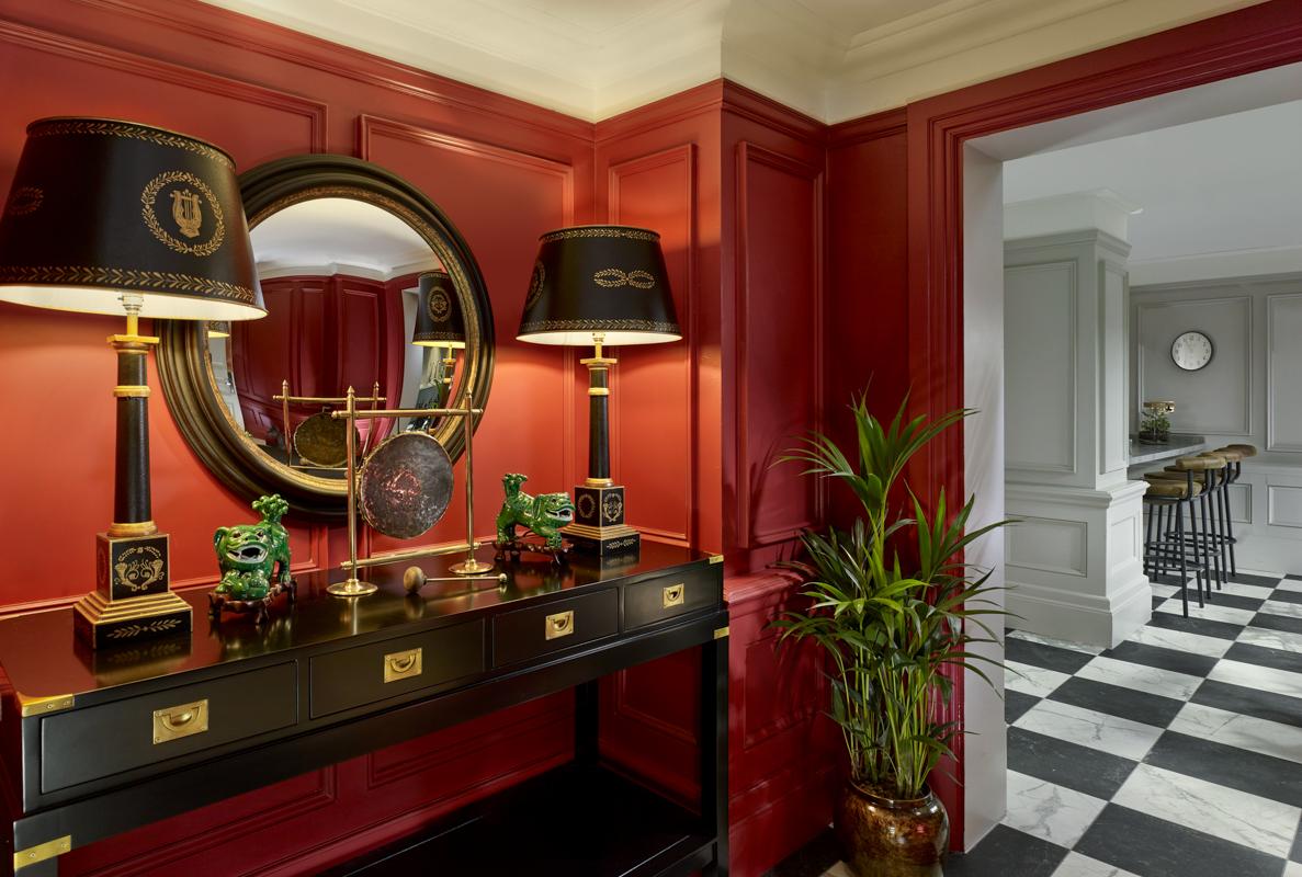 A hotel entry area with red paneled walls, a black console table, decorative table lamps, and a round mirror at The Residence at Holmes Hotel London.