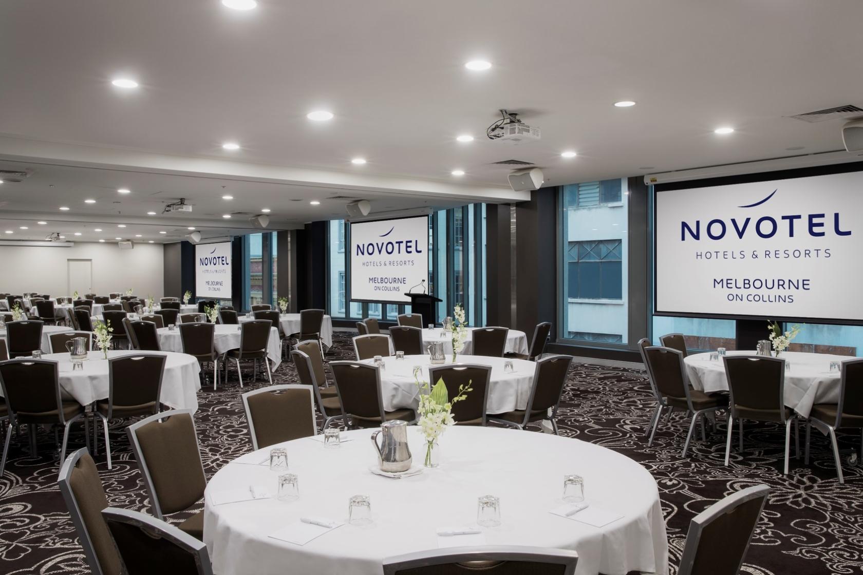 A function room with round tables, brown upholstered chairs, floral centerpieces, and floor-to-ceiling windows at Novotel Melbourne on Collins.