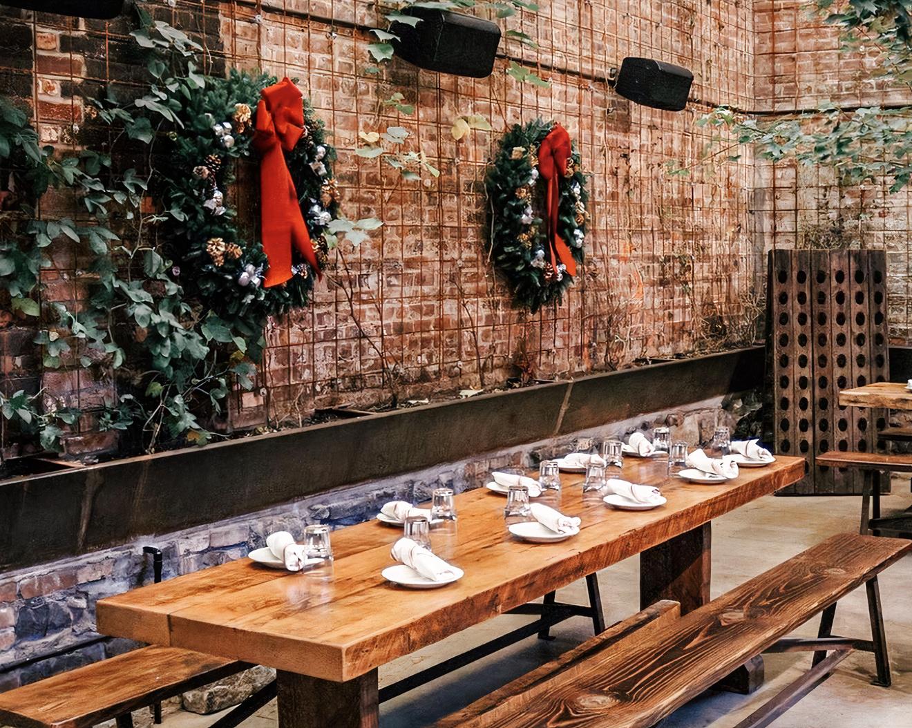 A function room with wood tables, exposed brick walls, and holiday wreaths at Ainslie Bowery.
