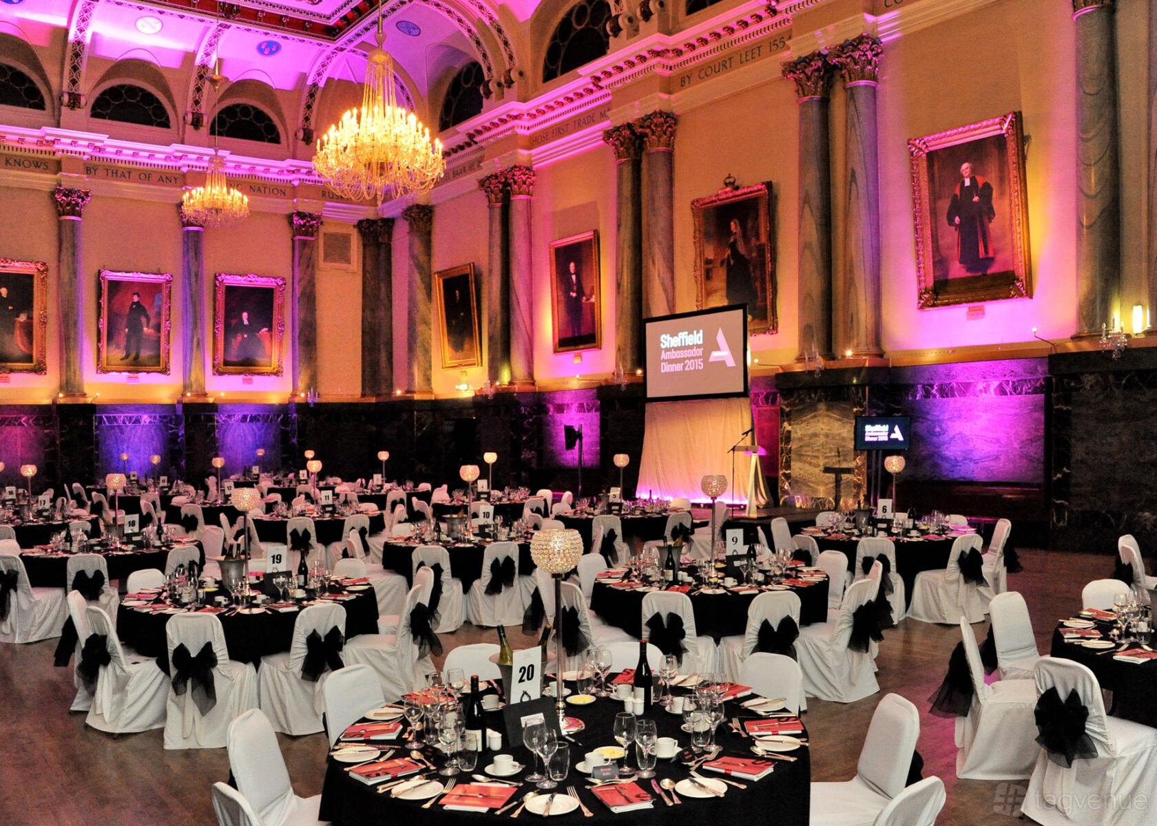An event hall with round tables covered in black and white linens, chandeliers, and uplighting at Cutlers’ Hall Hospitality.