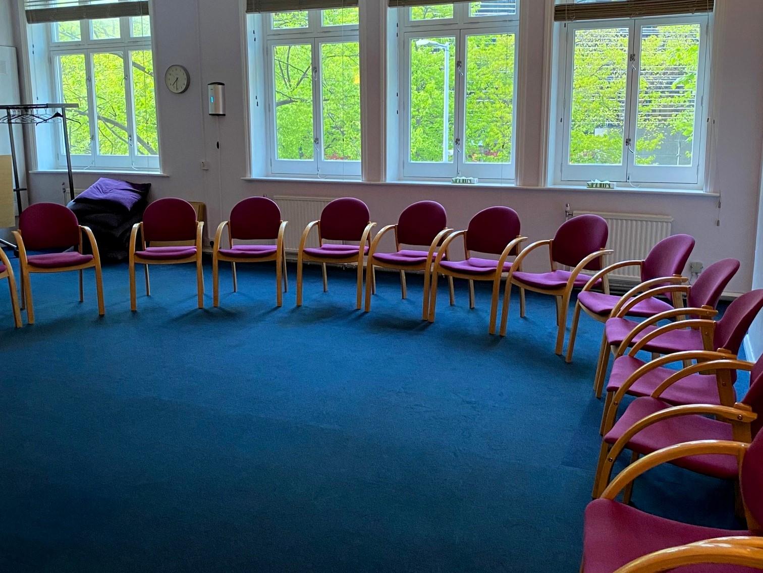 A meeting room with circular arrangement of red cushioned chairs and large windows at The Psychosynthesis Trust.
