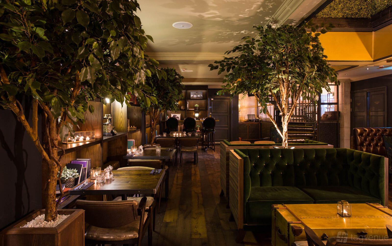 A bar with indoor trees, green velvet booths, wooden tables, and candlelit accents at The Lost and Found Greek Street.