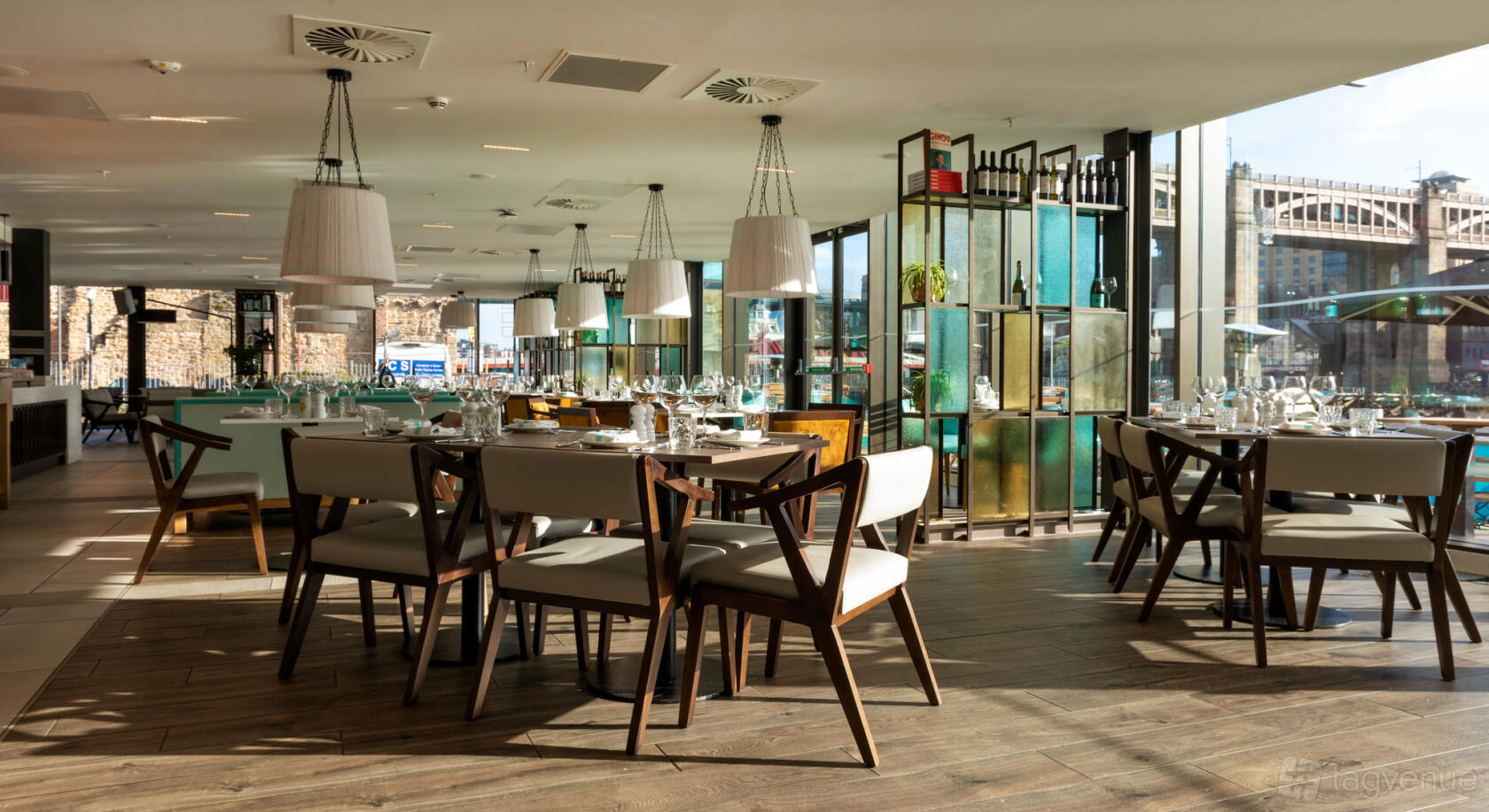 A restaurant with floor-to-ceiling windows, wooden floors, set dining tables, and pendant lighting at Gino D'Acampo Quayside.