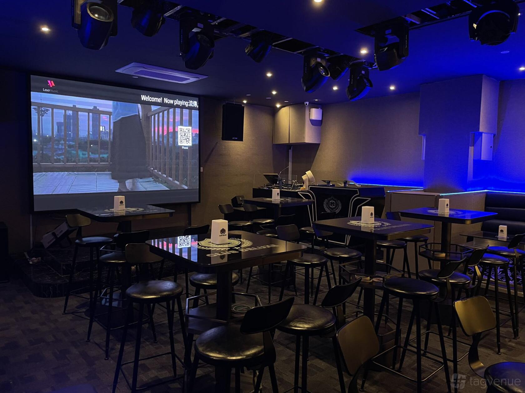 A nightclub with black high-top tables, bar stools, blue accent lighting, and a projection screen at Club Deluxe.