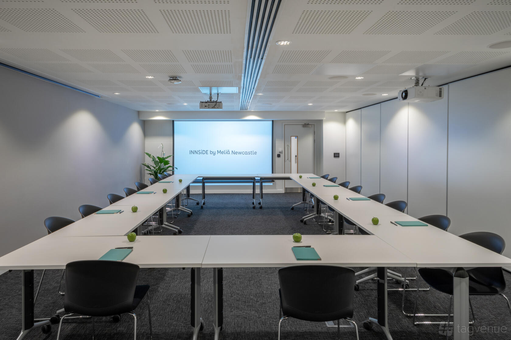 An event space with U-shaped tables, black chairs, a projector screen, and minimalist decor at INNSiDE By Melia Newcastle.
