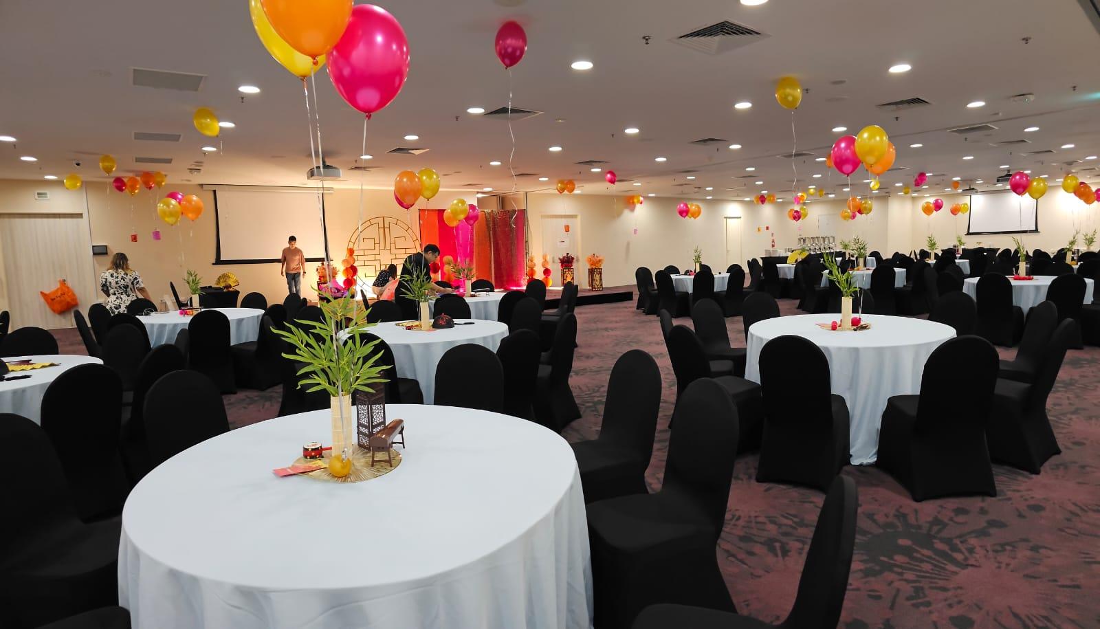 A hall with round tables dressed in white linens, black chairs, and colorful balloons at THE LEGACY, One-North.