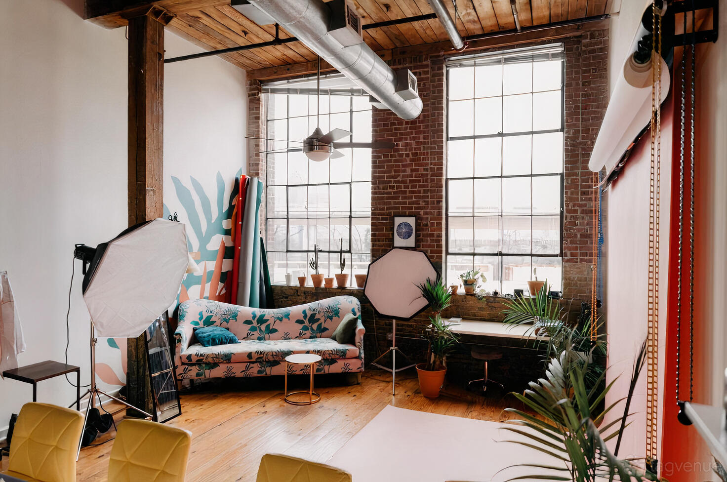 A photo studio with exposed brick walls, large industrial windows, photography lights, and a floral sofa at Eden Studios.