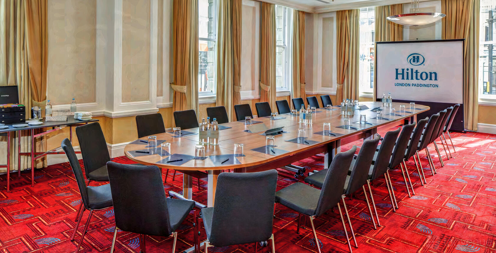 An auditorium with large windows, red carpet, U-shaped table setup, and presentation screen at Hilton London Paddington.