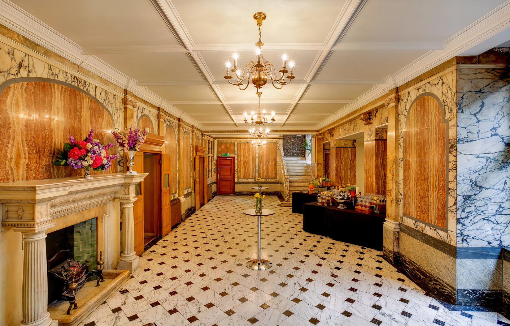 A hall with marble walls, chandeliers, a detailed fireplace, and flower arrangements at One Birdcage Walk.