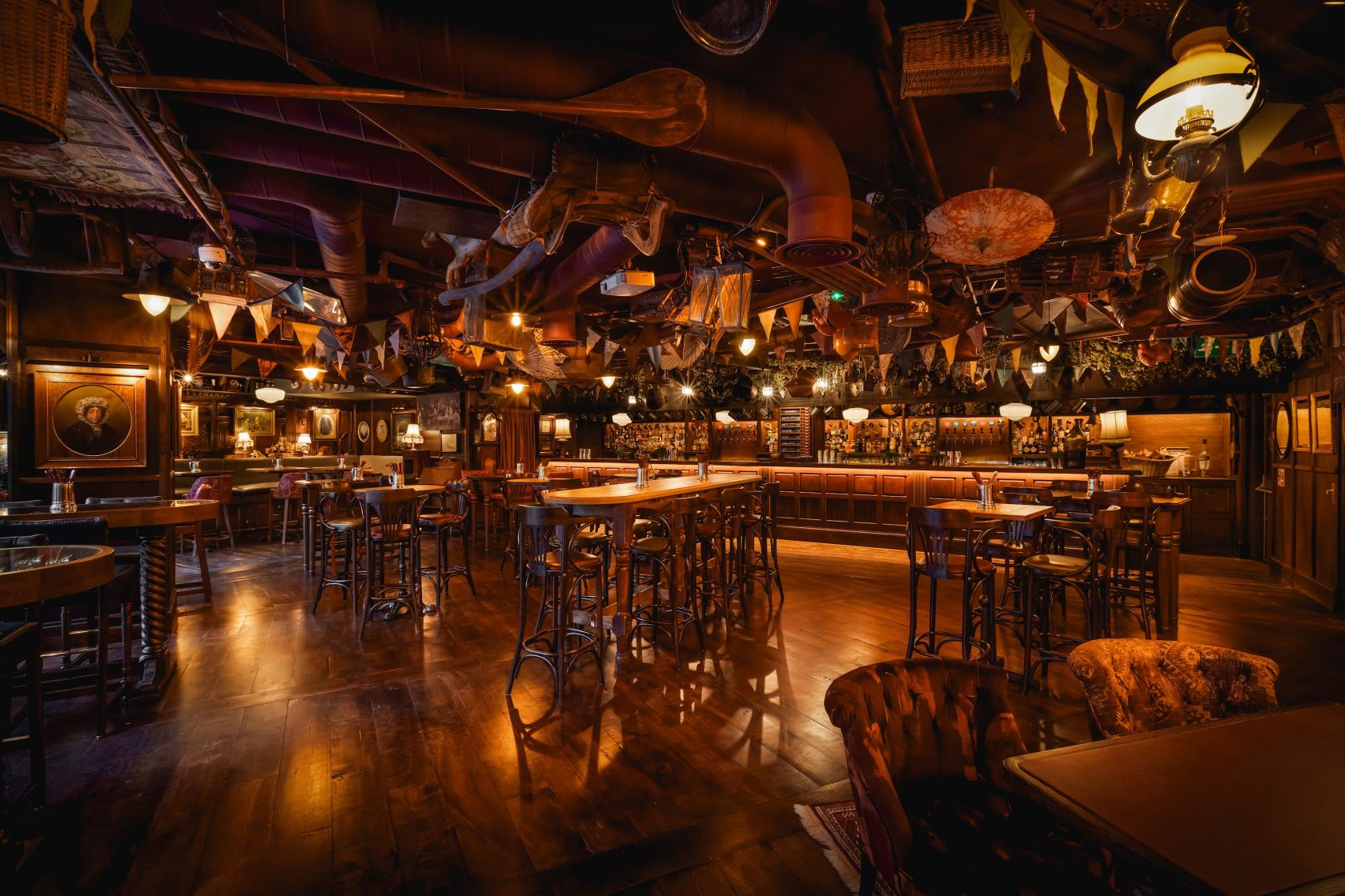 A cocktail bar with dark wood floors, vintage decor, flags, and high tables at Mr Fogg's City Tavern.
