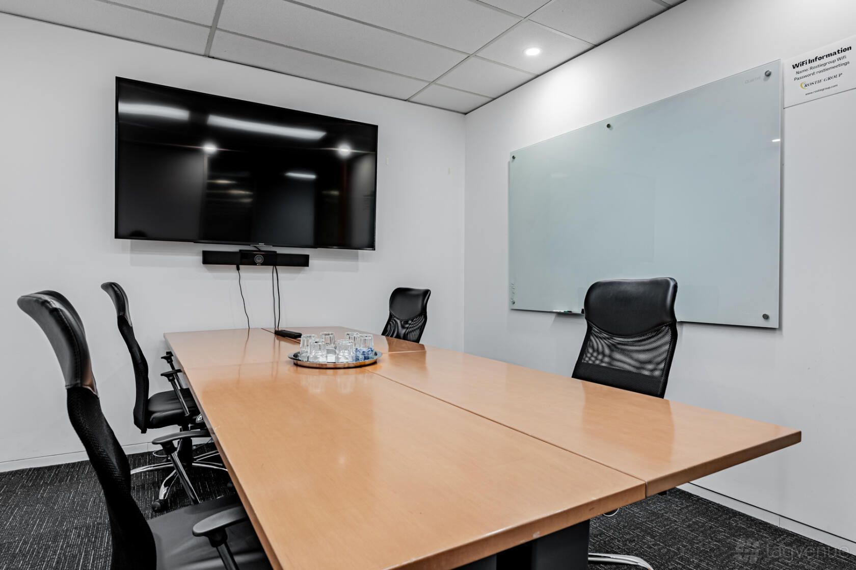 A meeting room with a wood conference table, black chairs, a wall-mounted screen, and a glass whiteboard at The Rostie Group.