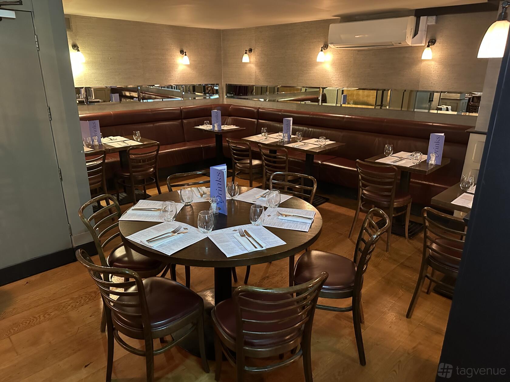 A restaurant with round wooden tables, brown leather banquette seating, and wall mirrors at Cote Brasserie Kensington.