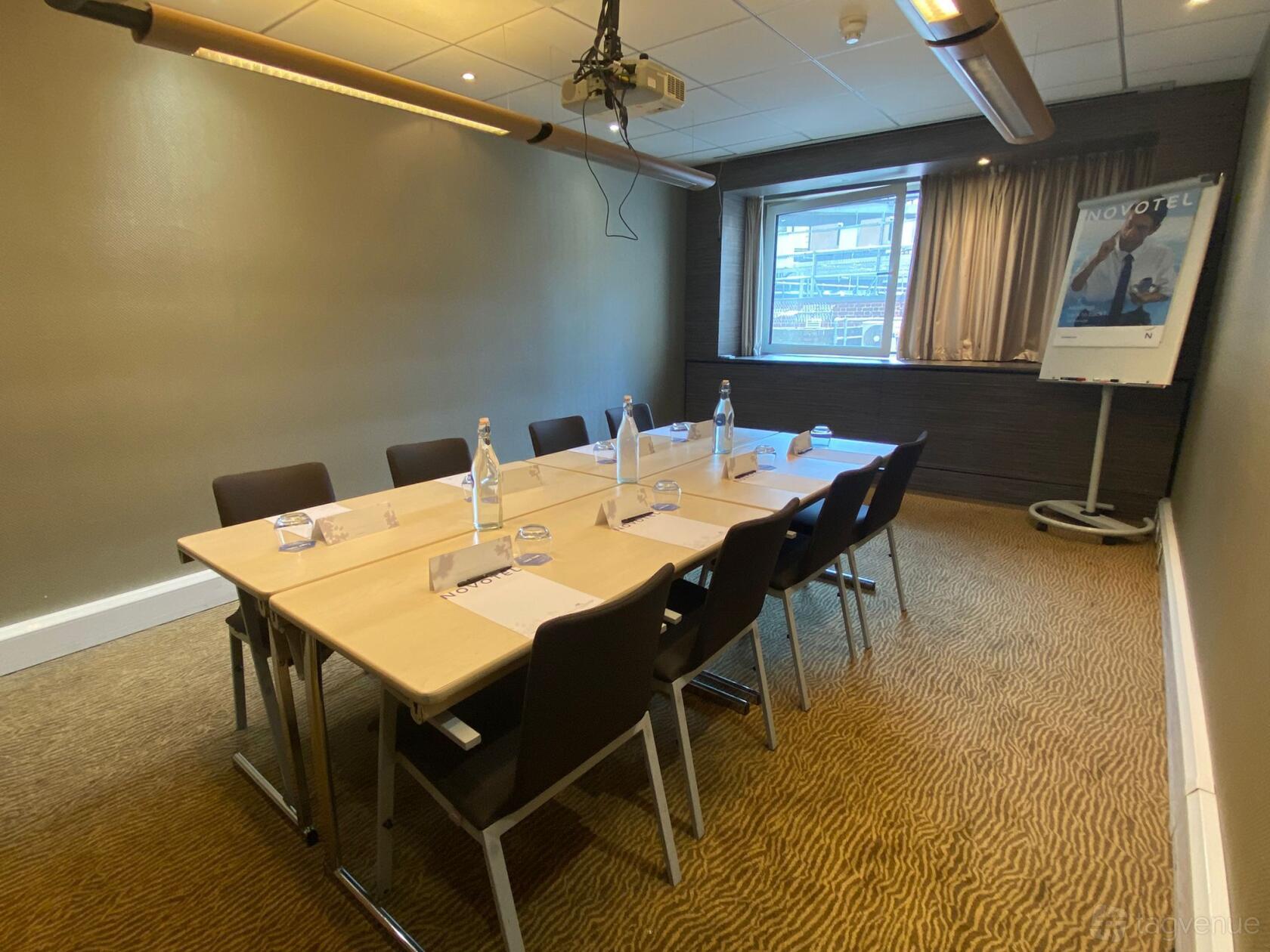A meeting room with rectangular tables arranged in a square, bottles of water, and a projector at Novotel London Waterloo.