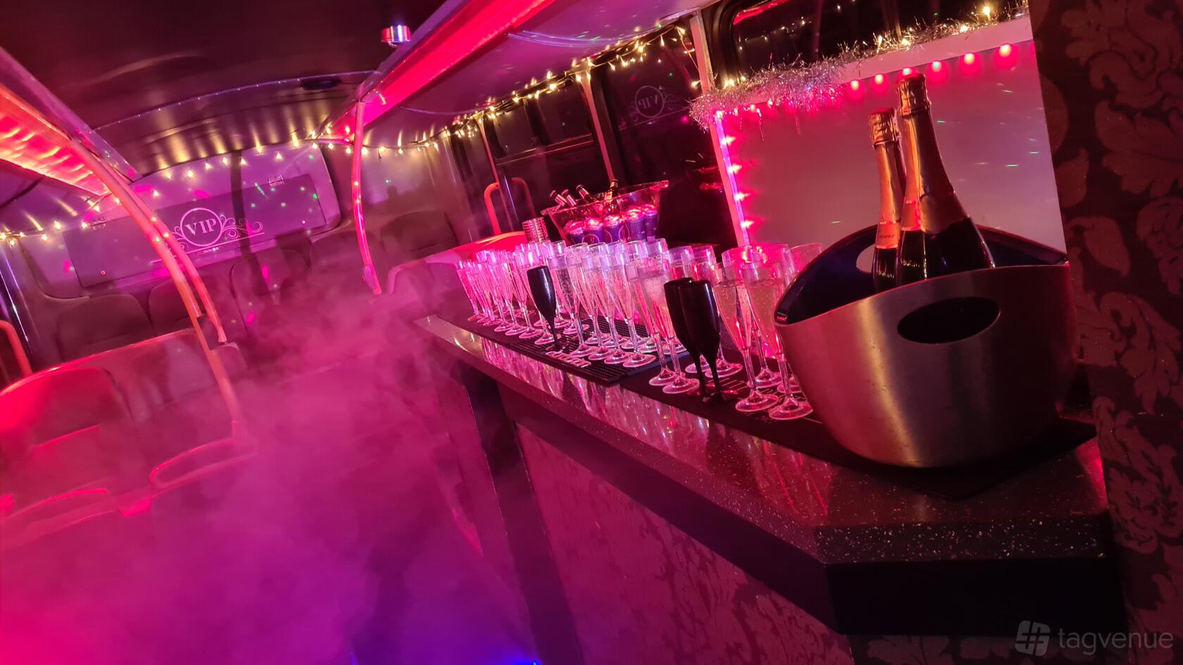 A party bus with champagne bottles and rows of filled glasses on a counter with pink lighting at Party Bus London.
