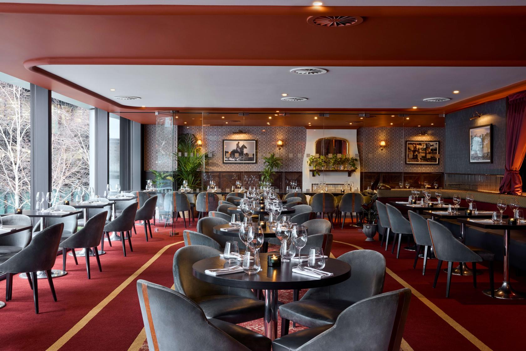 A dining room with floor-to-ceiling windows, round tables set with glassware, and red carpeting at Gaucho Tower Bridge.