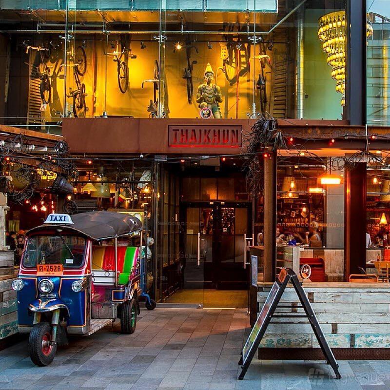 A restaurant entrance with floor-to-ceiling windows, a colorful tuk tuk taxi, and warm lighting at Thaikun Manchester.