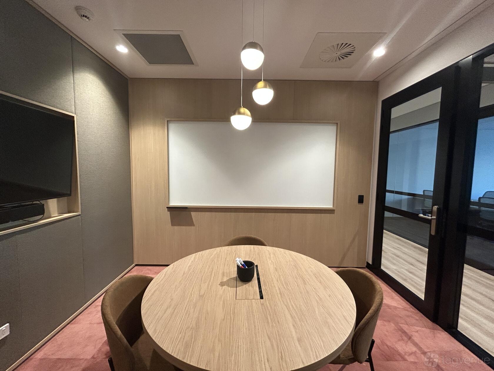 A meeting centre room with a round wooden table, wall-mounted whiteboard, pendant lights, and a wall display at JustCo at King Street.