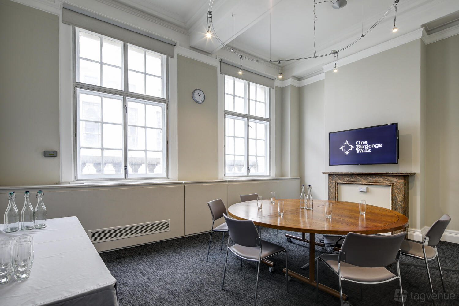 A meeting room with tall sash windows, a round wooden table, and a wall-mounted display at One Birdcage Walk.