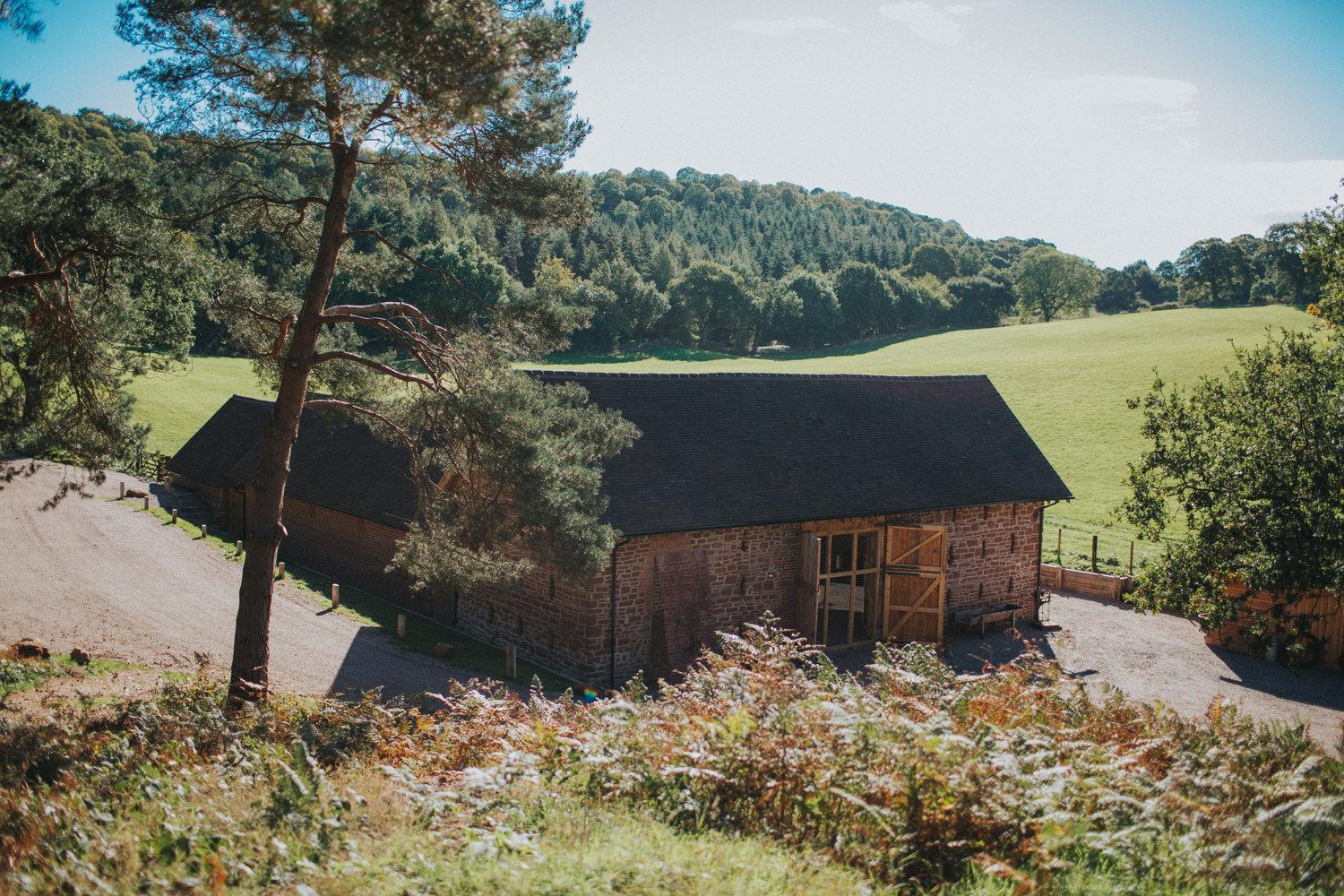 A rustic barn with wooden siding and large entry doors set beside open fields and surrounded by trees at Bridal Barn.