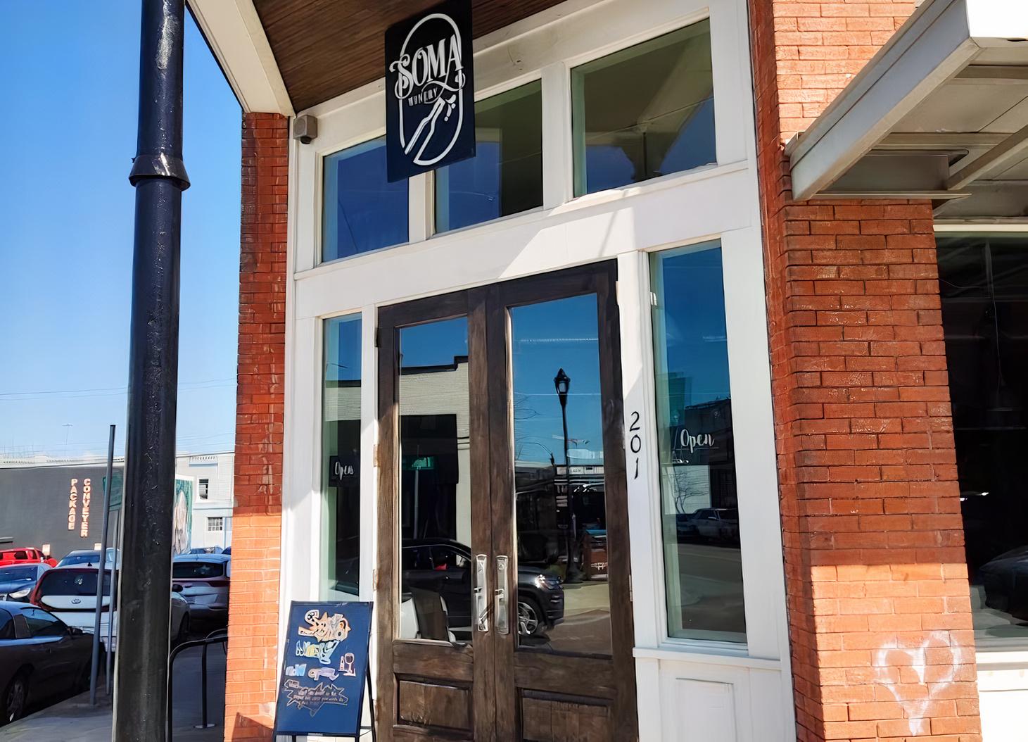 A pub entrance with large glass windows, double wooden doors, and a hanging sign at Soma Winery.