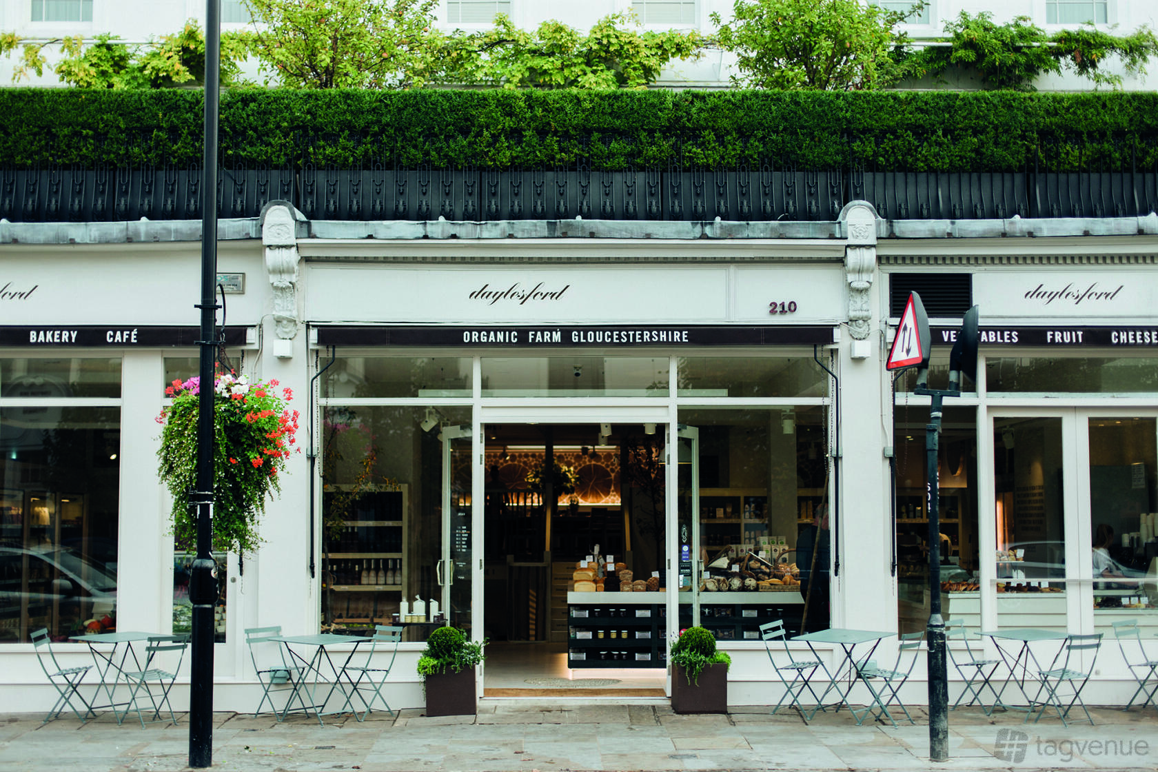 A café with outdoor seating, large front windows, and greenery above the entrance at Daylesford Organic Notting Hill.