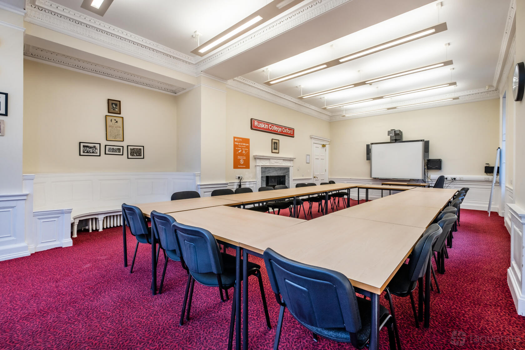 A boardroom with U-shaped tables, blue chairs, and a wall-mounted screen at Ruskin Venues.