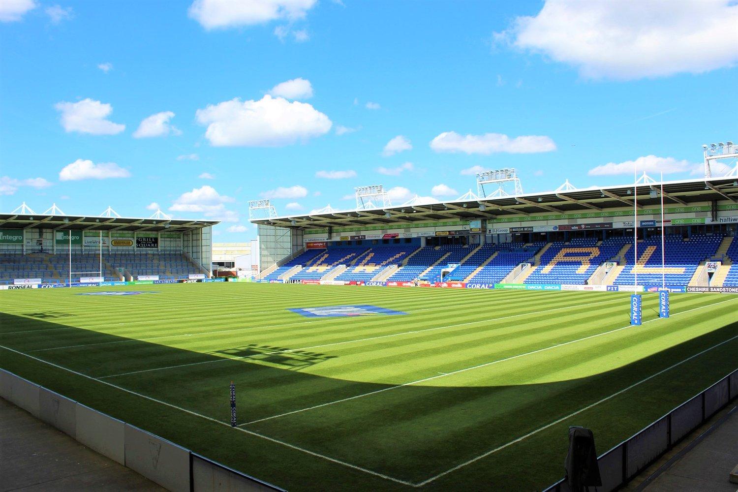 A stadium with tiered blue seating, a well-maintained grass pitch, and rugby posts at The Halliwell Jones Stadium.
