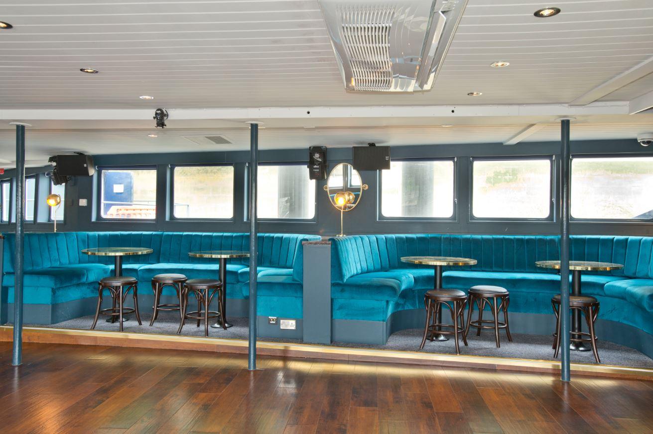 An event space on a boat with teal velvet banquette seating, round brass tables, and large windows at The Tattershall Castle.