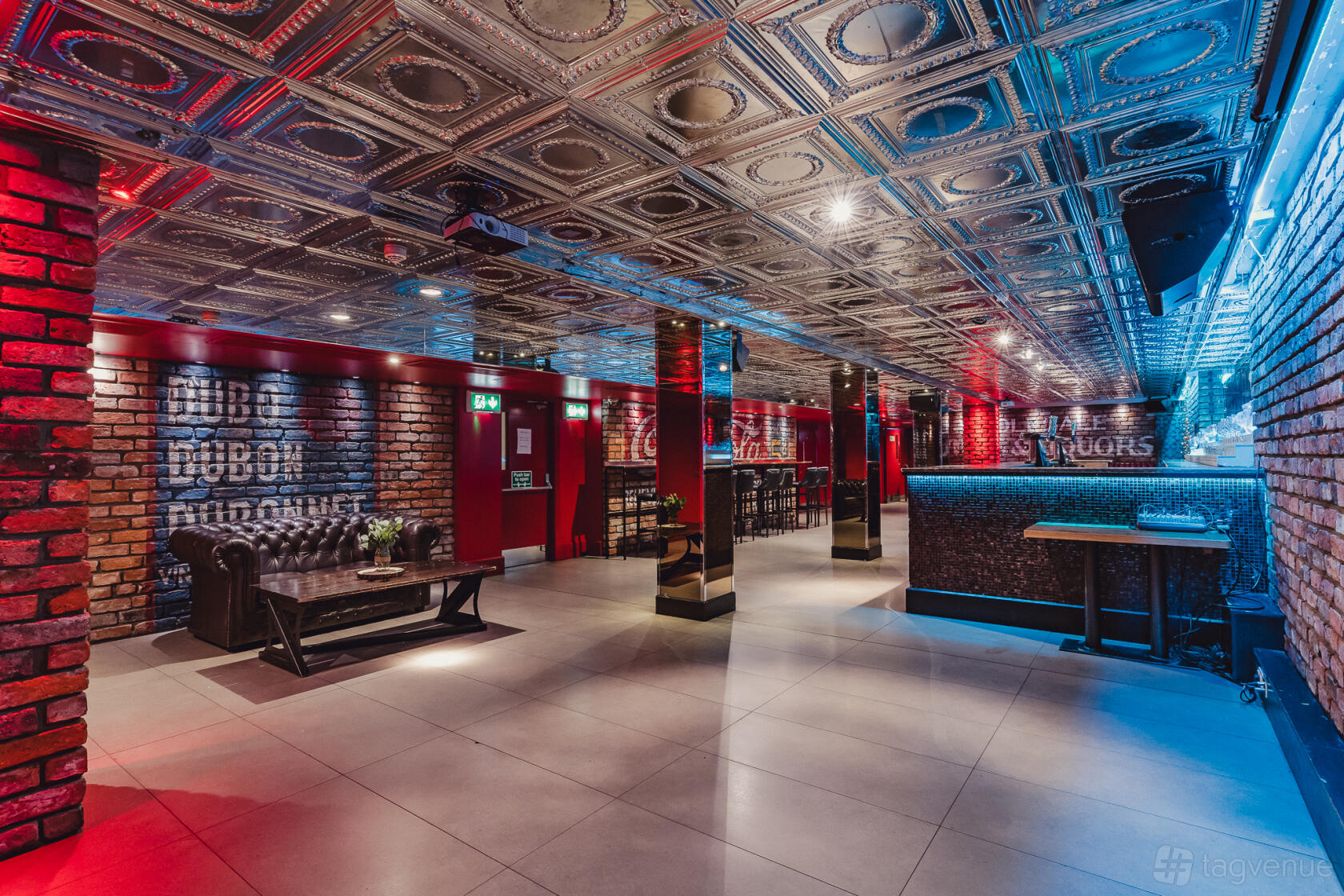 An underground space with exposed brick walls, ornate tin ceiling, and ambient blue-red lighting at Angel's Share Hotel.