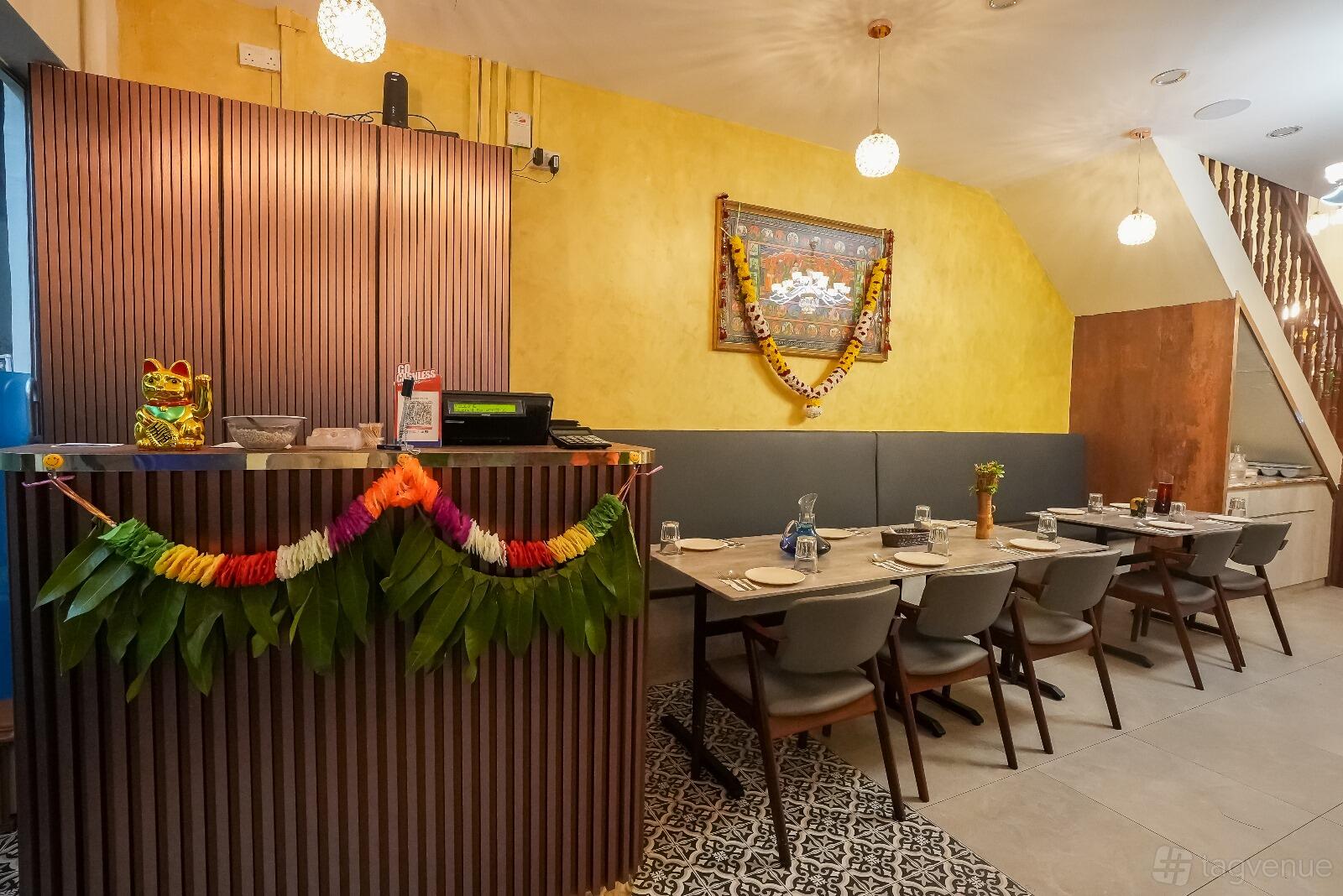 A restaurant with a long dining table, gray chairs, pendant lighting, and festive wall decorations at SARIGAMA Indian Restaurant.