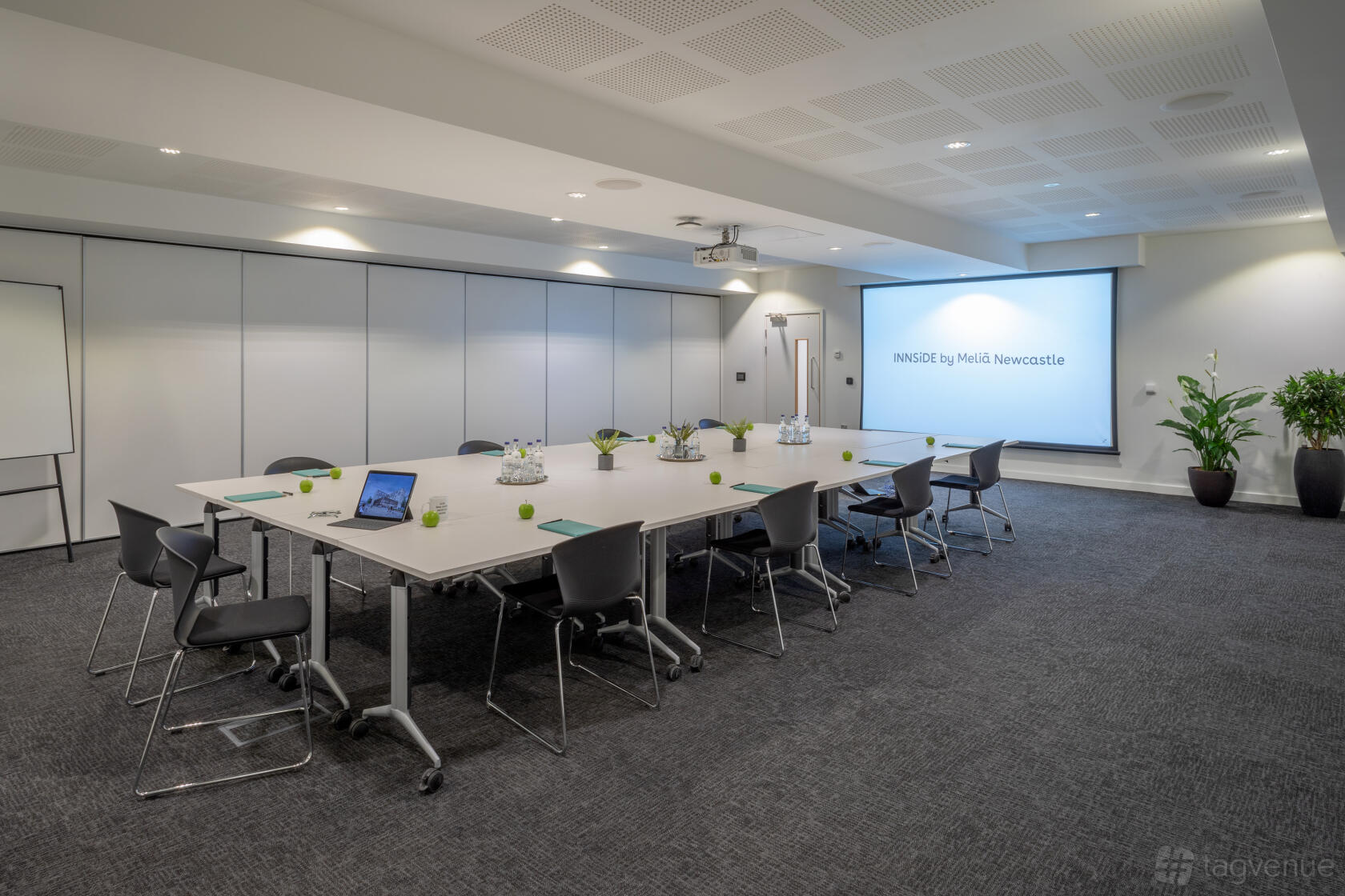 An event space with a large conference table, black chairs, a projector screen, and potted plants at INNSiDE By Melia Newcastle.