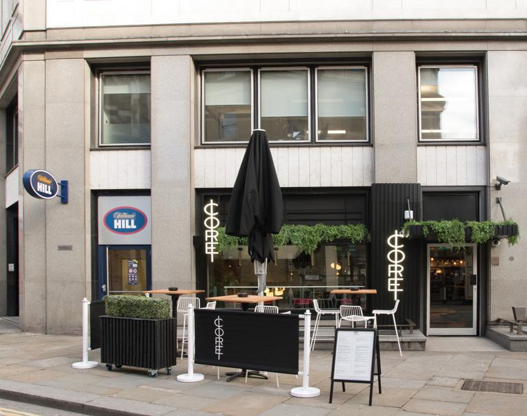 A pub with outdoor high tables, potted greenery, and black signage at Core.