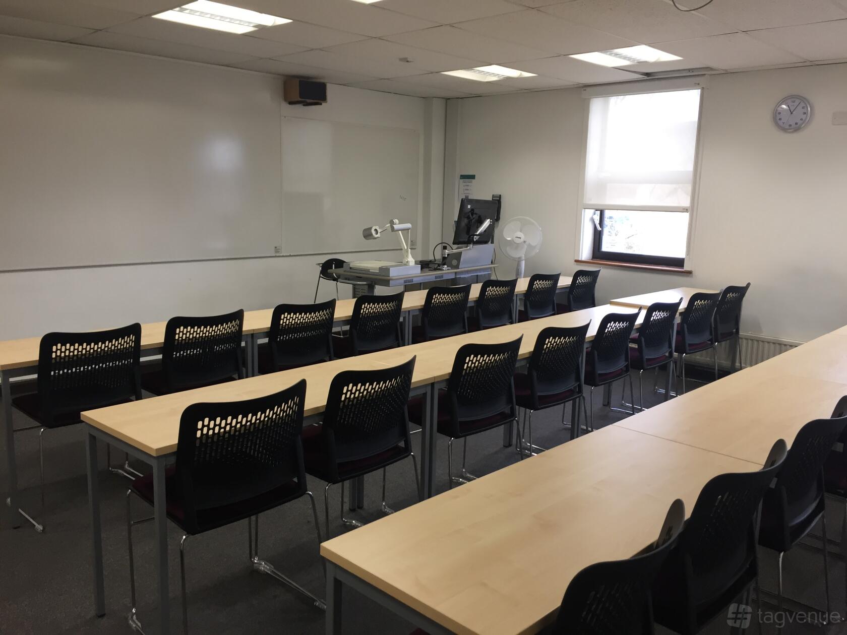 A classroom with rows of black chairs, light wood tables, and a whiteboard at ARU Venue Hire - Cambridge.